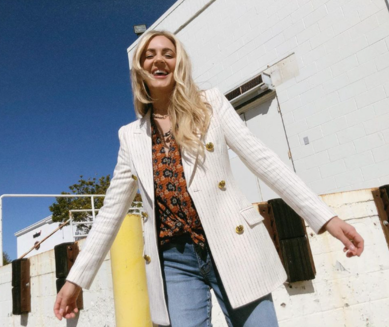 Blonde woman smiling in front of industrial building while wearing jeans, a orange and blue floral print blouse, and a white blazer.