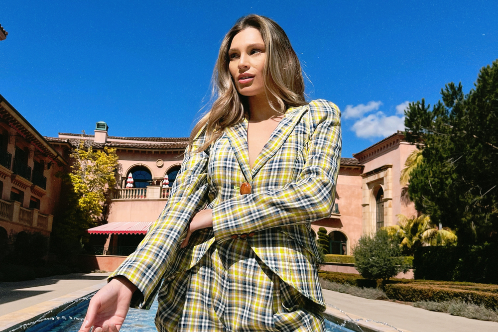Model in checkered suit coat and shorts standing in front of pool and rustic luxury building